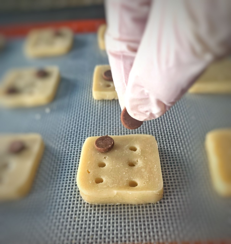 Image describing a batch of braille cookies to be baked, with a hand putting chocolate chip which represent braille dot on top of a cookie