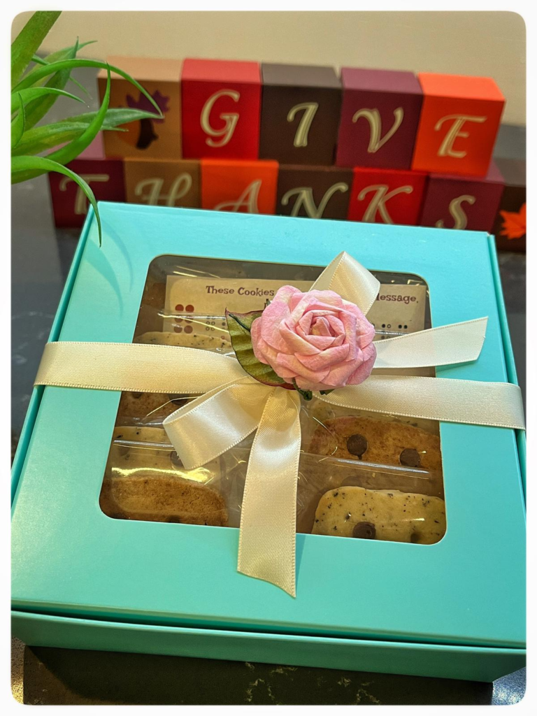 A blue box of braille cookies wrapped with a rose ribbon with a red and black wooden block behind reads "Give Thanks"