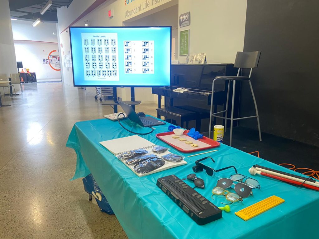 Image describing the reception desk of the braille cookie event with a monitor showing braille letters chart and some event related items: blind folder, special effect glasses, braille cookie making tray and braille letter equipment