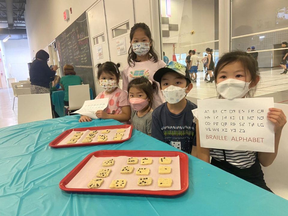 Image describing 5 kids standing behind the table with their braille cookie finished product and one kid holding the braille alphabet chart
