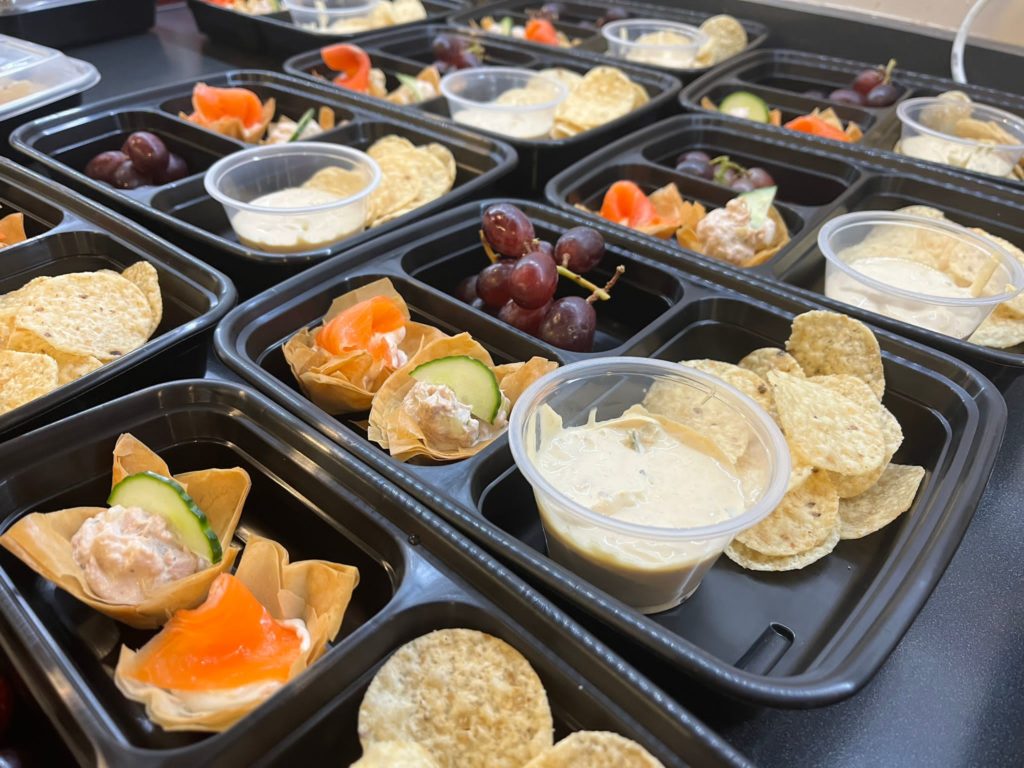 Image showing the snack boxes that are prepared by volunteers lining up on a table. In the box there are chips with dipping, grapes, and crispy smoked salmon appetizer.