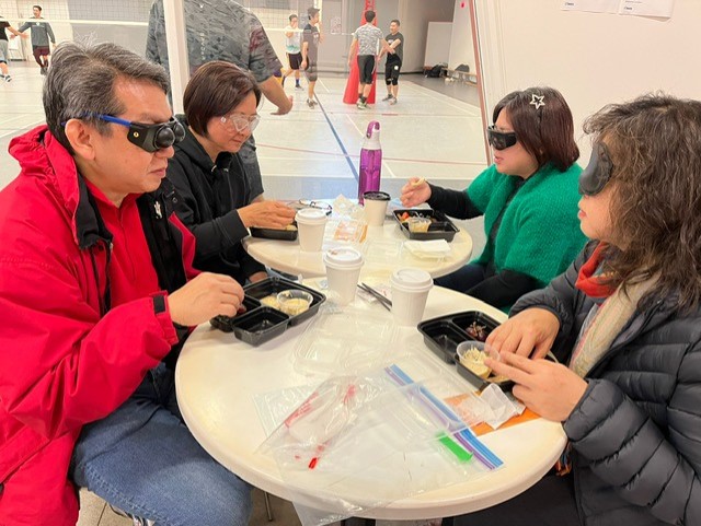 Image describing event participants grapping food from the snack box while wearing special glasses that simulate the effect of having various types of eye disease